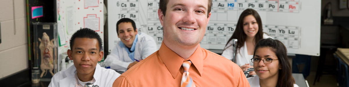 A smiling man in an orange shirt and patterned tie stands in front of four coworkers, with chemistry-style wall charts and diagrams in the background.