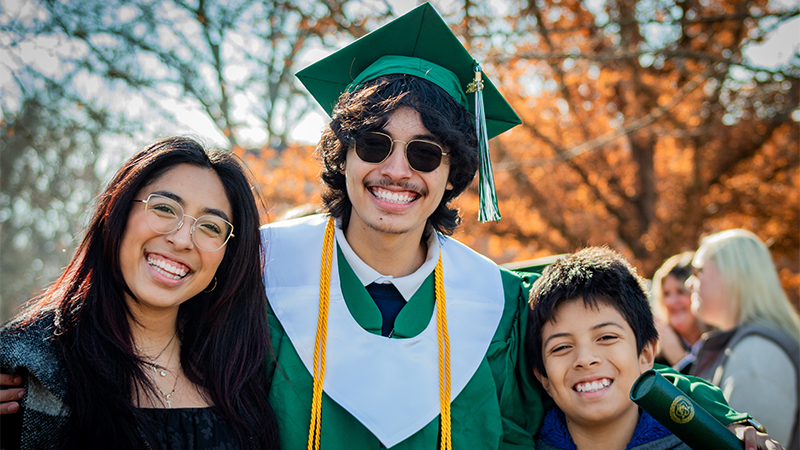A young man in a green graduation cap and gown stands smiling in between his sister and brother with his arms wrapped around them.