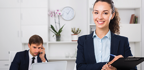 A woman with a clipboard standing in front of a man on a computer