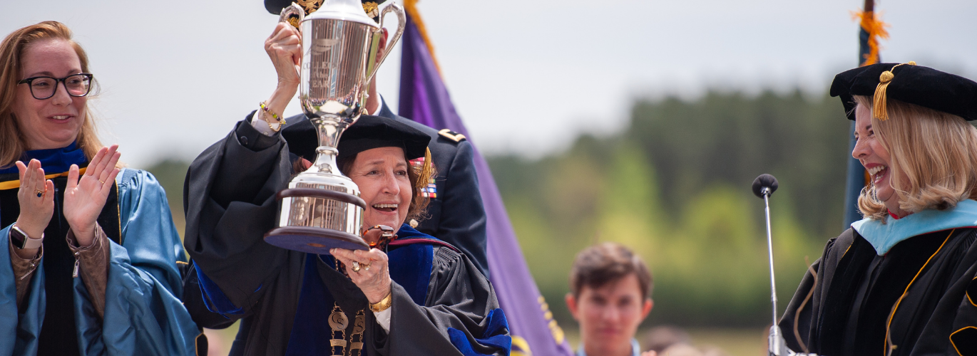 Janet Smith stands on stage in regalia holding a large trophy with Flora Tydings in regalia