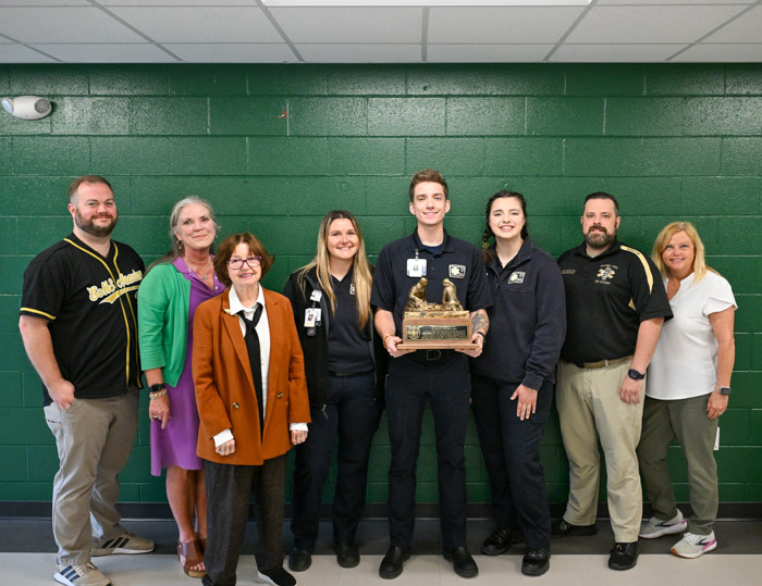 A group of people stand in front of a dark green wall while holding a large wood and metal trophy.