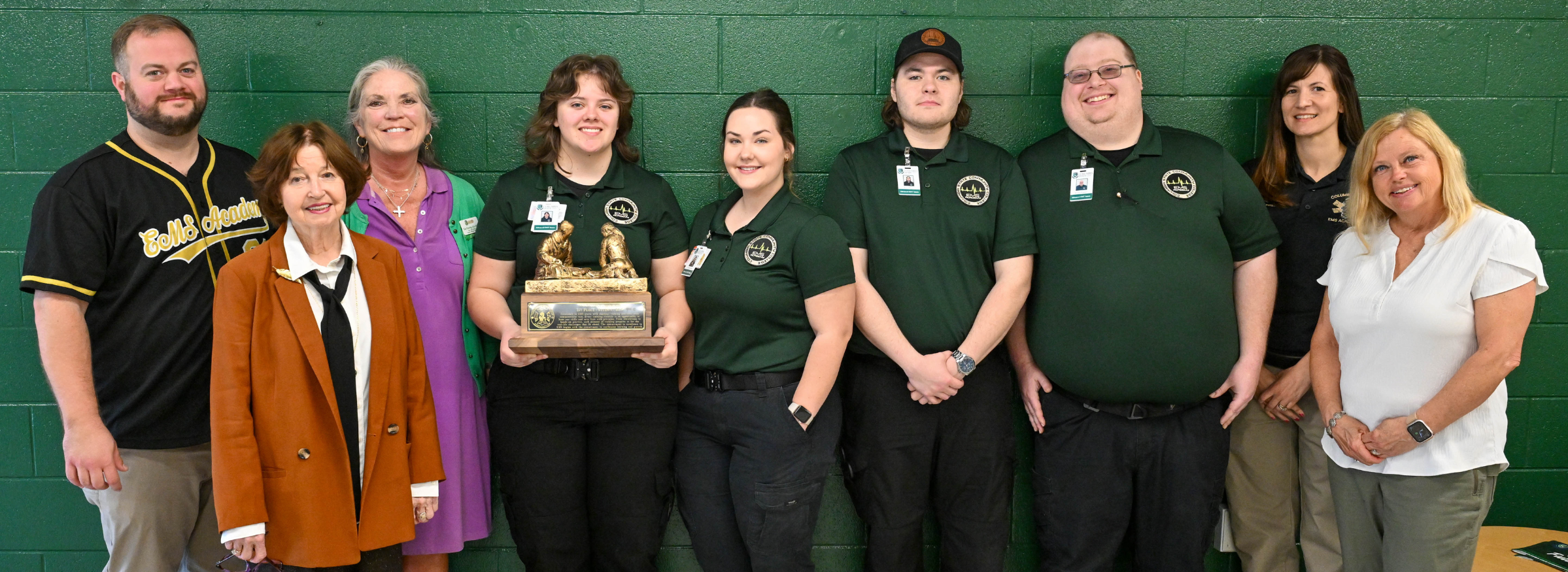 A group of people stand in front of a dark green wall while holding a large wood and metal trophy.