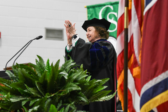 A woman in graduation gown claps while standing at a podium.