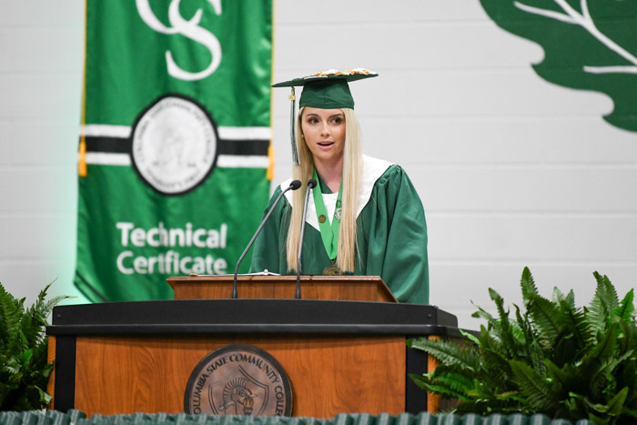 A person in a green cap and gown speaks at a podium.