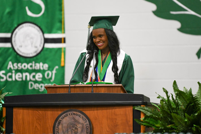 A person in a green cap and gown speaks at a podium.