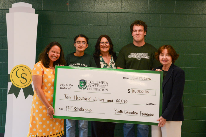 A group of people stand in front of a dark green wall holding a large check.