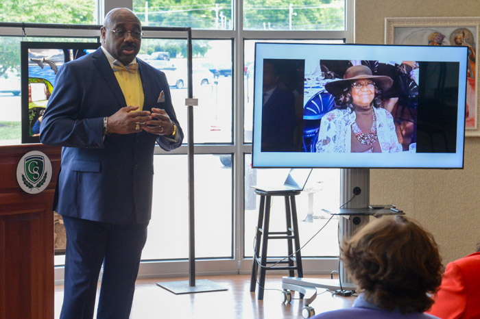 A person stands talking to a room of people while a television monitor shows a picture of an older woman.