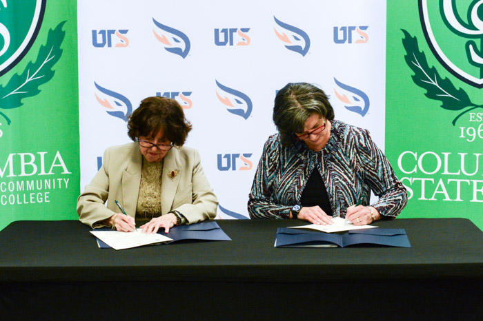 Two people sit at a table and sign sheets of papers in folders.