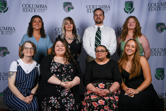 Eight people sit and stand in front of a background. 