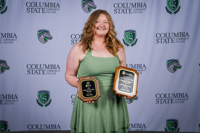 A person stands in front of a white background while holding two wooden award plaques.