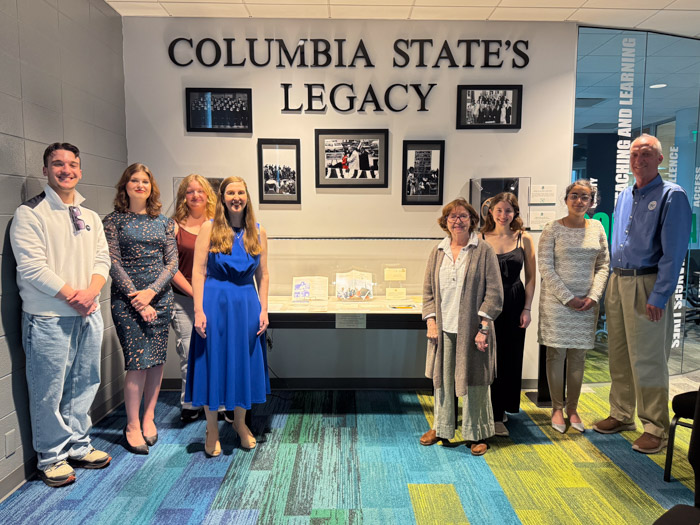 A group of people stand in front of a wall with display cases and framed pictures with a large sign that says "Columbia State's Legacy."
