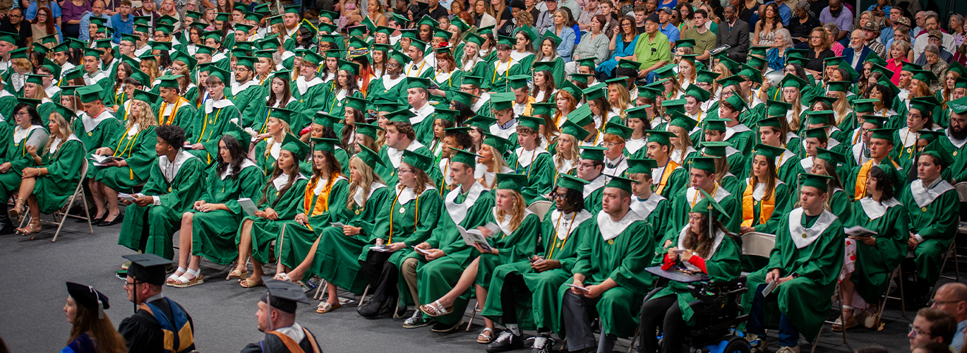 A wide-shot photo shows a large group of people sitting in green caps and gowns.