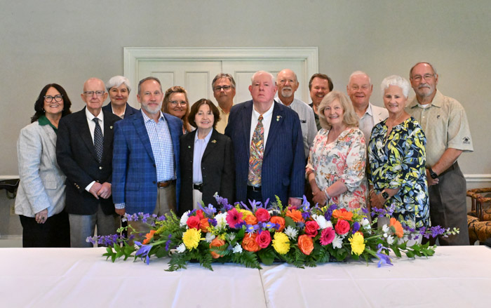 A large group of people stand in two rows behind a bouquet of flowers on a table.