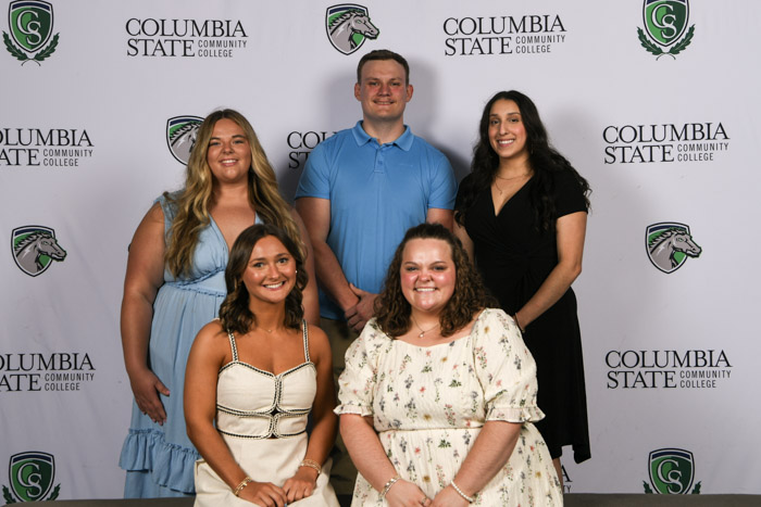 A group of students wearing professional attire both sit and stand for a photo in front of a white background.