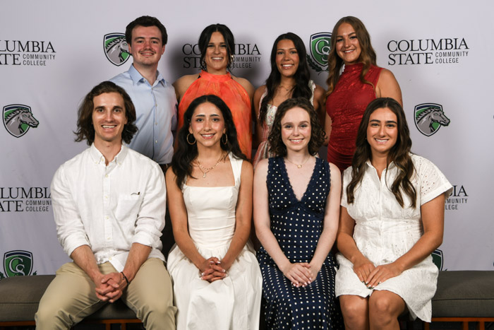 A group of students wearing professional attire both sit and stand for a photo in front of a white background.