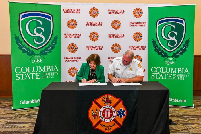 Two people sit at a table and sign sheets of papers in folders with white and green backgrounds behind them.