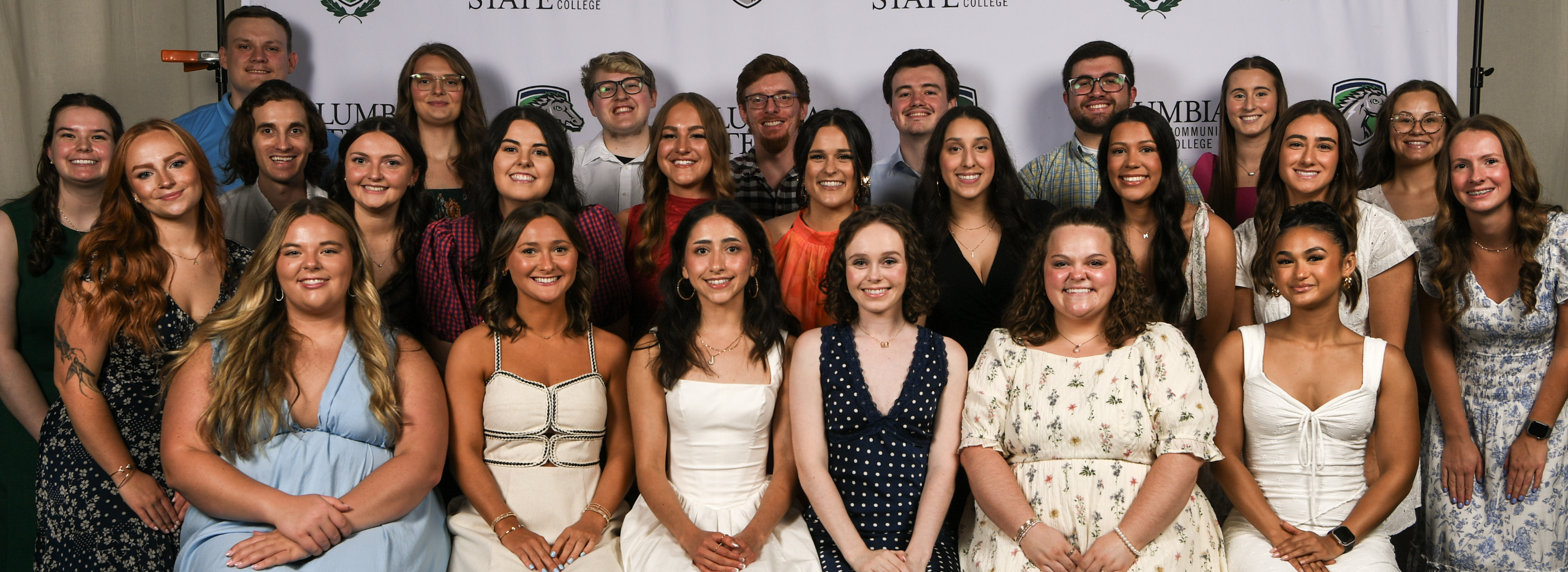 A photo shows a group of people wearing professional clothing both standing and sitting in front of a white background.