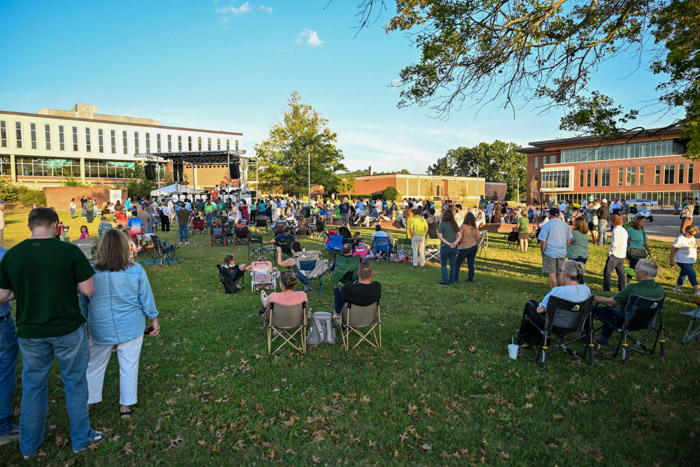 A photo shows a wide shot of people standing and sitting in a grassy area facing towards a stage.