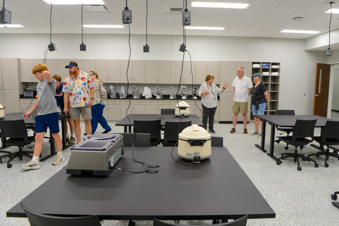 People stand and walk through a lab classroom with microscopes and machines on tables. Electric outlets hand from the ceiling.