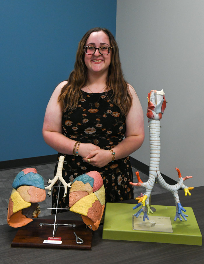A person stands and smiles for a photo with two anatomical displays of lungs and bronchioles in front of them.