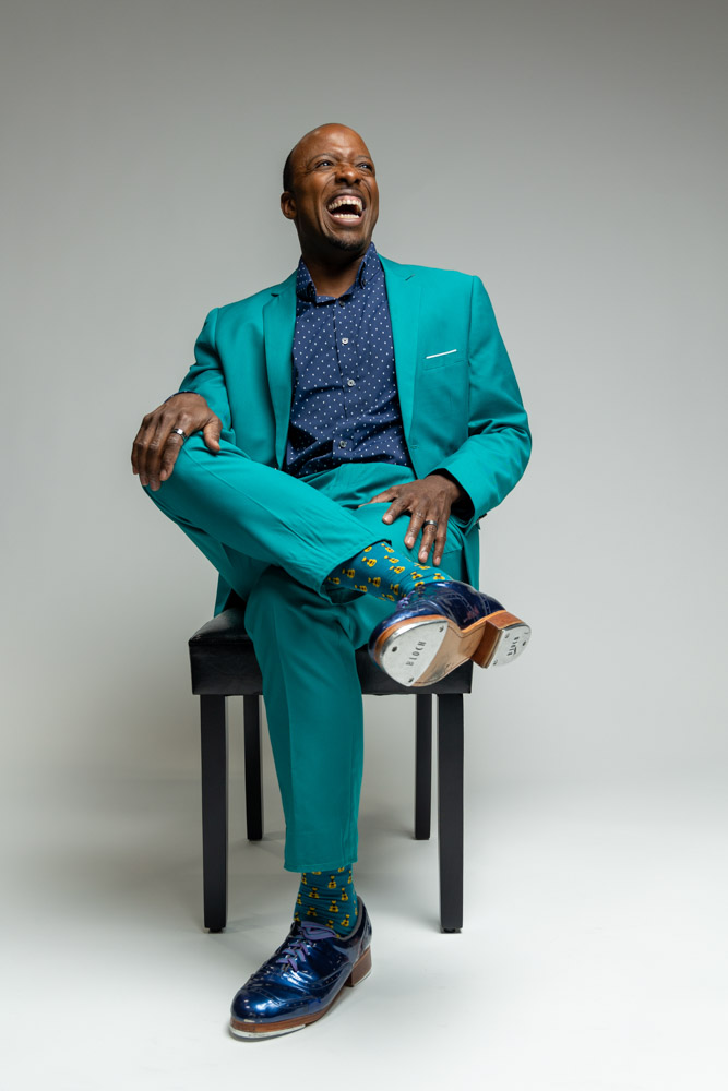 A man in a teal suit and tap shoes smiles while seated.