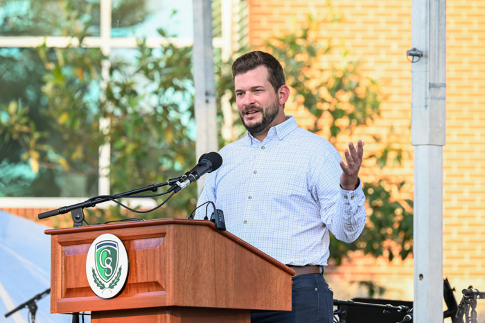 A person wearing a white and blue shirt speaks at a wooden podium.