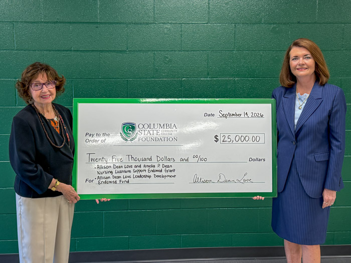 Two women on opposite sides hold a large signed check with a dark green wall behind them.