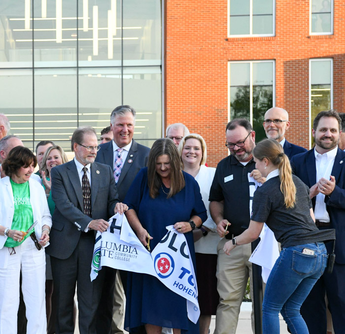 A group of people standing in a group in front of a building all cut through a large white ribbon banner.