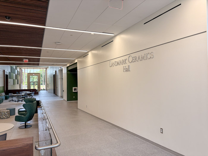 A long hallways leading to an entrance with windows has gray tile floors and white walls with the words "Landmark Ceramics Hall."