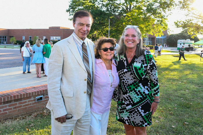 Three people wearing formal attire stand and smile outside for a photo.
